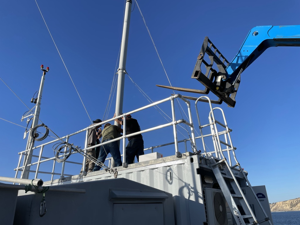 Technicians hold up the inlet stack for installation as part of the Aerosol Observing System. A piece of the ocean appears to the right.