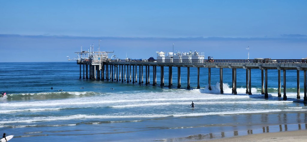 Surfers and swimmers frolic in the water around the Scripps Pier in La Jolla, California. A line of five instrument containers sits along the pier with a cloud bank visible on the horizon.
