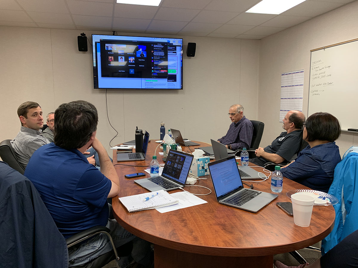 Six people sit around a conference table during a workshop, with a large screen showing those attending virtually.