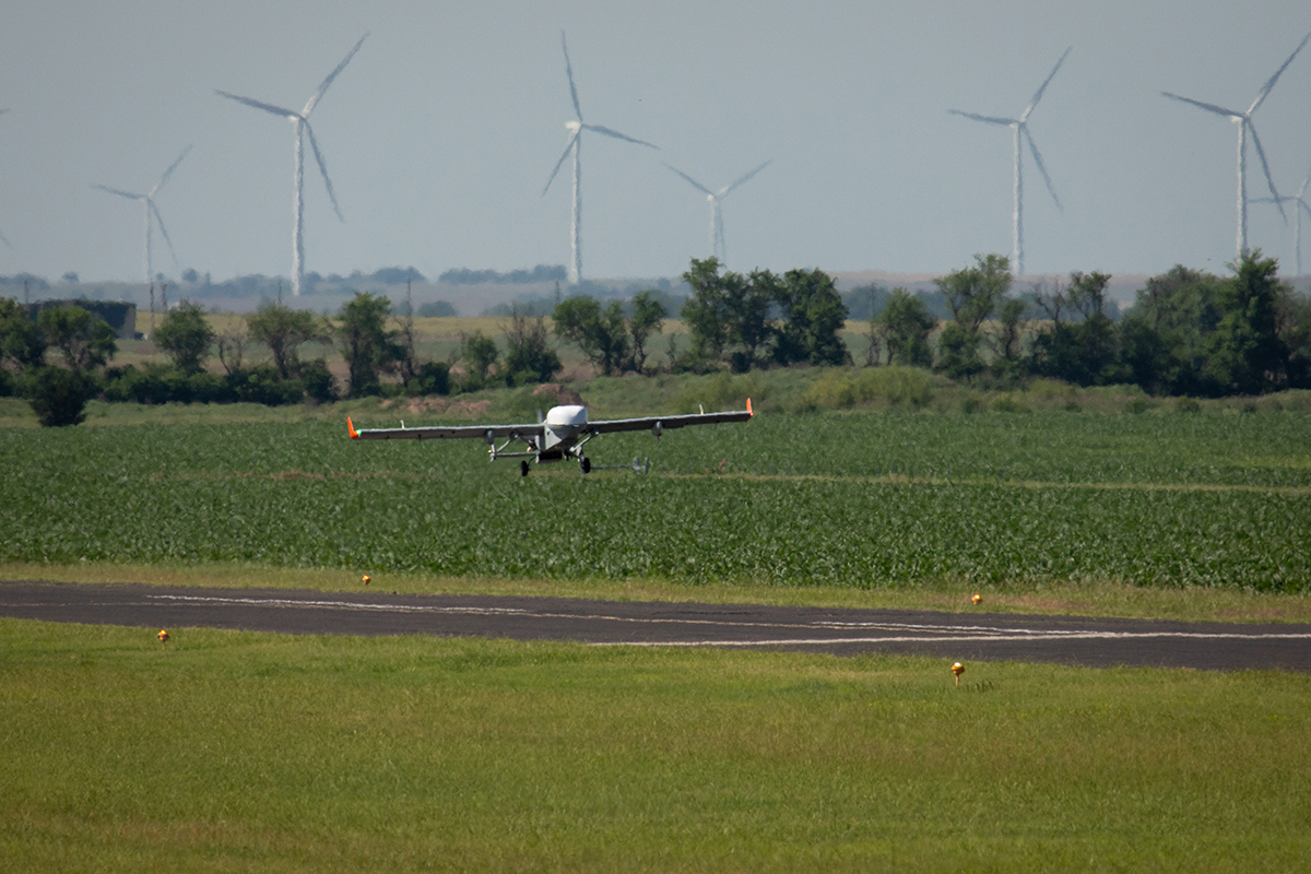 With wind turbines in the distance, the ArcticShark uncrewed aerial system is captured over the runway at the Blackwell-Tonkawa Municipal Airport.