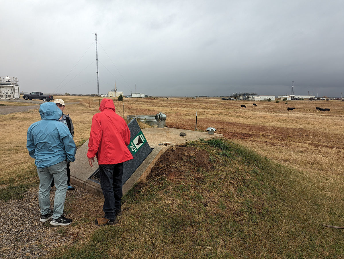 With clouds, cows, and atmospheric instruments as a backdrop, one person holds open a door to look inside a severe storm shelter while two others talk.